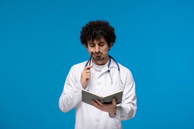 Arzttag, lockiger, hübscher, süßer Kerl in medizinischer Uniform, der einen Stift und ein Buch hält und denkt