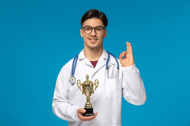 Arzttag süßer junger gutaussehender mann im laborkittel und brille, der ok-zeichen zeigt und trophäe hält
