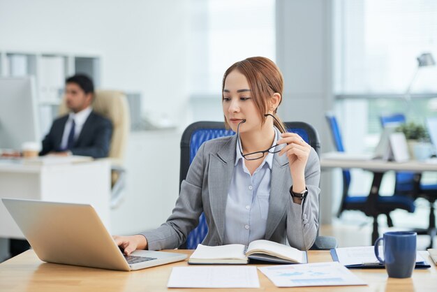 Asiatin, die am Schreibtisch im Büro sitzt, Gläser hält und an Laptop arbeitet