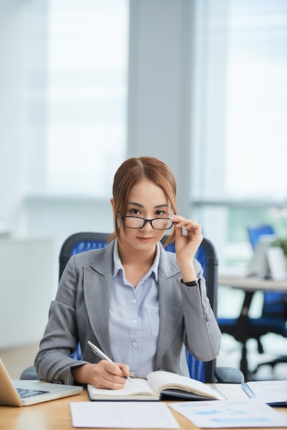 Asiatin, die am Schreibtisch im Büro sitzt, in Planer schreibt und nach vorn über Gläsern schaut