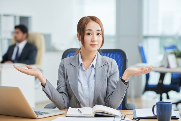 Asiatin, die am Schreibtisch im Büro sitzt und Kamera mit hilflosem Handzeichen betrachtet