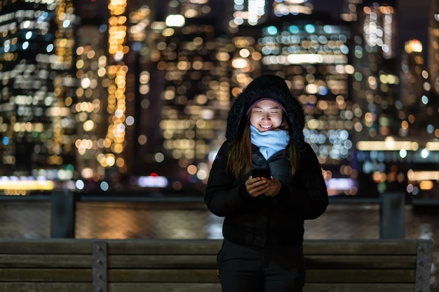 Asiatin in der winterklage unter verwendung des intelligenten handys mit lächelnaktion über dem foto verwischte bokeh