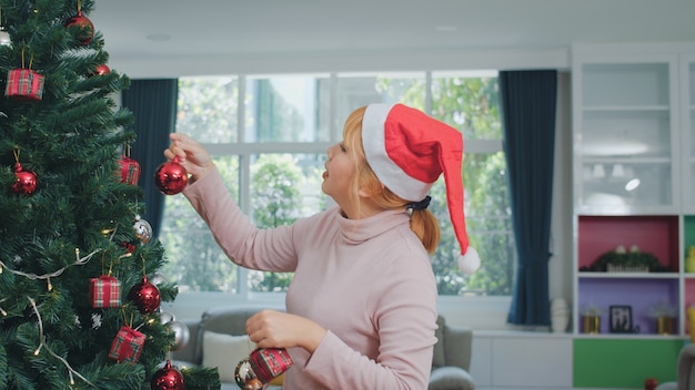 Asiatinnen verzieren Weihnachtsbaum am Weihnachtsfest. Das weibliche jugendlich glückliche Lächeln feiern Weihnachtswinterferien im Wohnzimmer zu Hause.