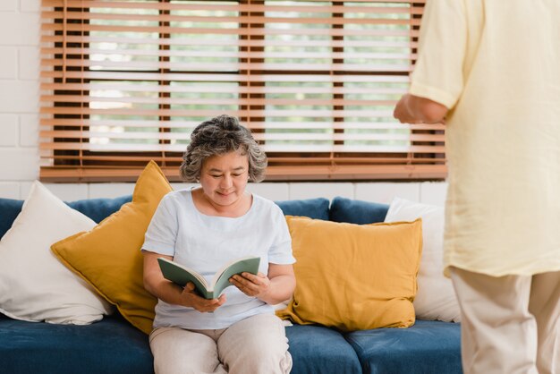 Asiatische ältere Frau, die zu Hause ein Buch im Wohnzimmer liest. Chinesisches weibliches Lügen auf Sofa, wenn Sie zu Hause entspannt werden.