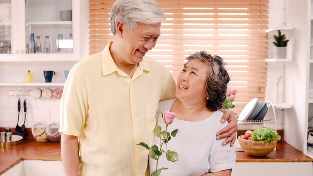 Asiatische ältere Paare, die dem glücklichen Lächeln und Halten der Blume und Schauen zur Kamera sich fühlen, während zu Hause in der Küche sich entspannt. Lebensstil-ältere Familie genießen Konzept der Zeit zu Hause. Porträt, Blick in die Kamera.