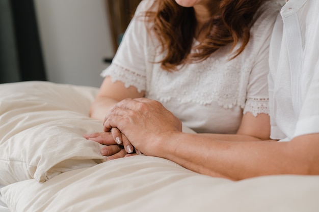 Asiatische ältere Paare, die ihre Hände beim Schlafzimmer zusammennehmen, Paargefühl glücklich teilen und das Lügen auf Bett zu Hause stützen. Älteres Konzept der Familie des Lebensstils zu Hause.