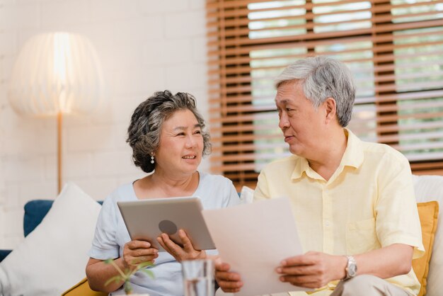 Asiatische ältere Paare unter Verwendung der Tablette, die zu Hause im Wohnzimmer fernsieht, Paare genießen Liebesmoment beim auf Sofa liegen, wenn Sie zu Hause entspannt werden.