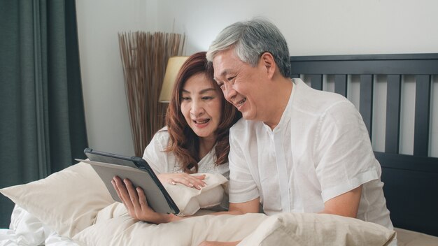 Asiatische ältere Paare unter Verwendung der Tablette zu Hause. Asiatische ältere chinesische Großeltern, Videoanruf, der mit Familienenkelkind spricht, scherzt beim auf Bett im Konzept des Schlafzimmers zu Hause morgens liegen.