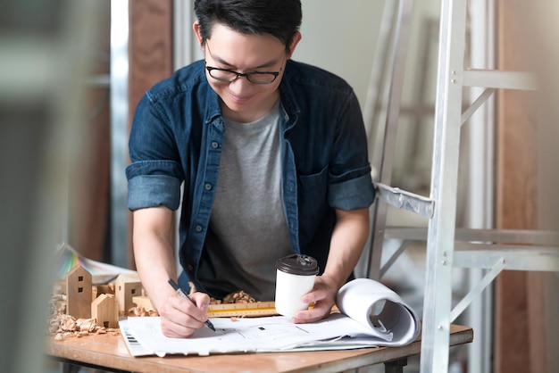 Asiatische attraktive Innenarchitektin mit Brille, die am Baufortschritt der Hausrenovierung mit Lächeln und Selbstvertrauen am Schreibtisch mit Blaupause und Zeichenwerkzeug arbeitet