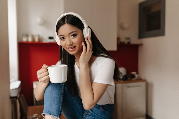 Asiatische braunäugige Frau, die Tasse Tee auf Hintergrund der Küche hält