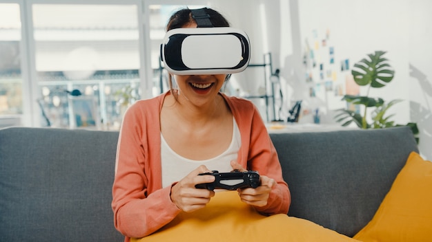 asiatische Dame tragen Headset-Brille der virtuellen Realität spielen Joystick-Spiel auf der Couch im Wohnzimmer im Haus