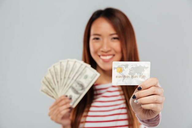 Asiatische Dame über grauer Wand, die Geld und Kreditkarte hält.