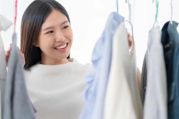 Asiatische frau des hübschen glücks, die mit dem spaß einkauft, der tuchkleid im weißen raumhintergrund des lagers auswählt