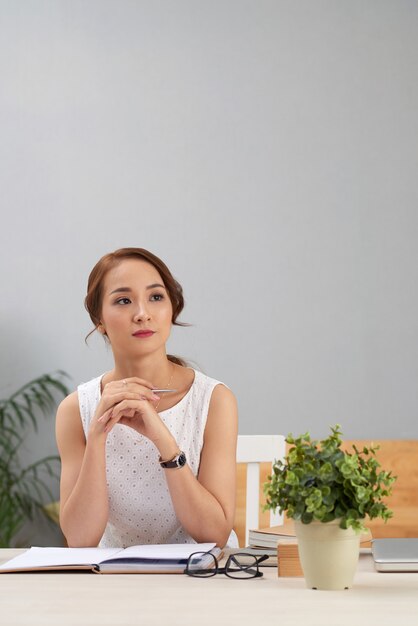 Asiatische Frau, die am Schreibtisch mit Zeitschrift sitzt, weg schaut und denkt