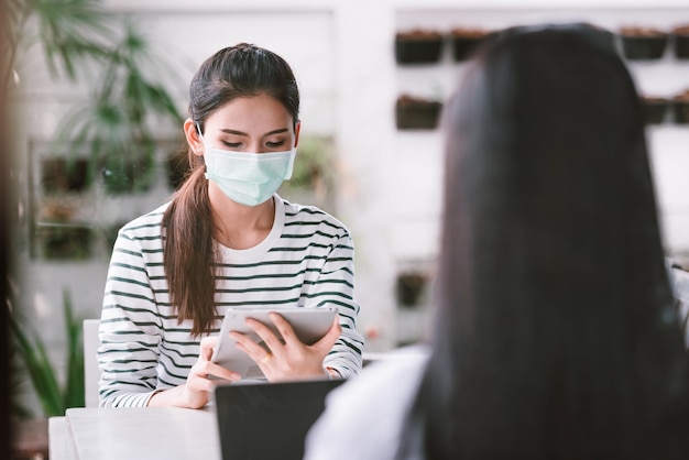 Asiatische Frau, die an einem Tablet arbeitet, während sie eine medizinische Maske im Café trägt Neue normale soziale Distanzierung des Lebensstils