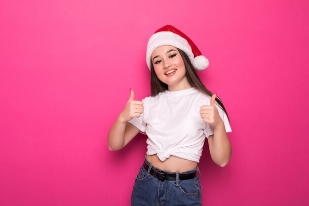 Asiatische Frau, die den Hut des Weihnachtsmanns mit den Daumen oben trägt, lokalisiert auf rosa Wand