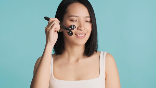 Asiatische Frau, die die Augen geschlossen hält, führt eine morgendliche Hautpflegeroutine durch und massiert das Gesicht mit einer Walze, die auf buntem Hintergrund isoliert ist