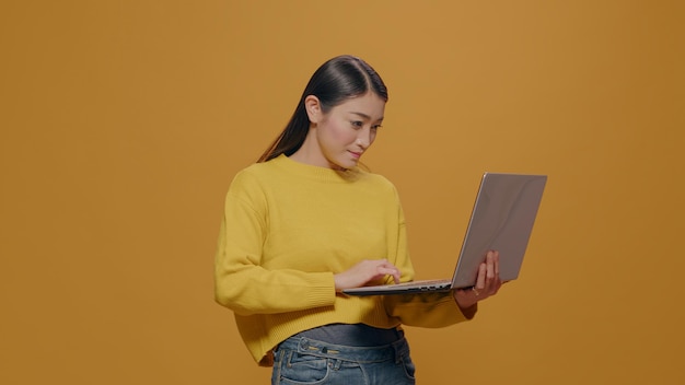 Asiatische frau, die laptop vor der kamera über gelbem hintergrund verwendet, computer hält und im internet surft, um an projekt zu arbeiten. kreativer unternehmer, der den gerätebildschirm im studio betrachtet. Kostenlose Fotos