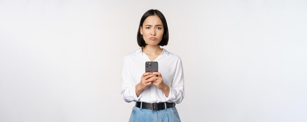 Asiatische Frau, die Smartphone hält und mit Zweifel enttäuscht von der Handy-App aussieht, die ag steht
