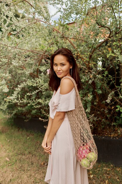 Asiatische Frau im Kleid, die umweltfreundliche Netzkäufertasche mit frischen tropischen Früchten hält.