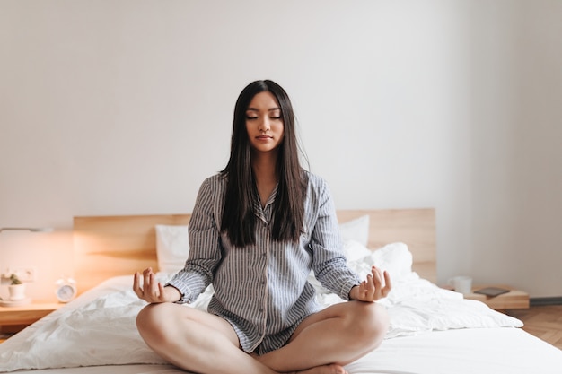 Asiatische Frau im Sommerpyjama meditiert sitzend auf weißem Bett