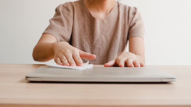 Asiatische Frau mit Alkoholspray auf Taschentuch sauber Laptop vor dem Öffnen zum Schutz Coronavirus. Weiblicher sauberer Computer für Hygiene, wenn soziale Distanzierung zu Hause bleibt und Selbstquarantänezeit.