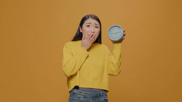 Asiatische Frau mit Angst, die die Zeit auf der Uhr im Studio überprüft, zu spät zur Arbeit kommt und in Verspätung ist. Ernsthafte Person, die auf Stunden und Minuten schaut und sich Sorgen um Fristen und schlechtes Timing macht.