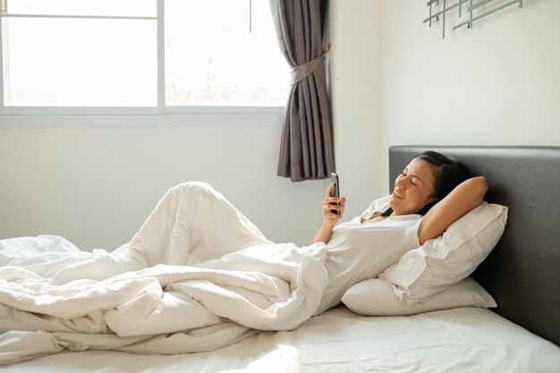 Asiatische Frau mit attraktivem Lächeln verwenden Tablet-Smartphone auf weißem Bett.