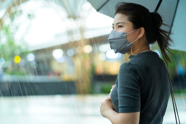 Asiatische Frau mit Gesichtsmaskenschutz, die in der Regenzeit bei starkem Regen im Freien auf der Straßenseite allein steht