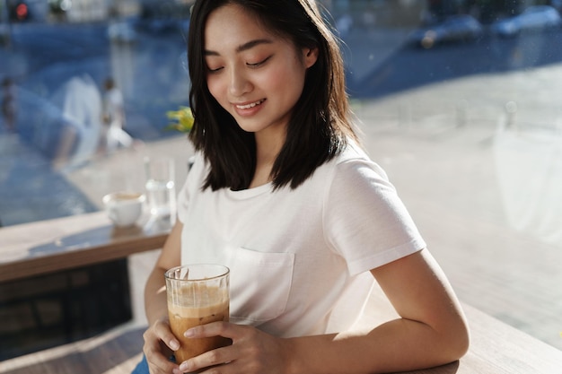 Asiatische Frau trinkt Eislatte im Café in der Nähe des Fensters und kühlt sich an heißen Sommertagen mit einem erfrischenden Getränk ab, das glücklich lächelt