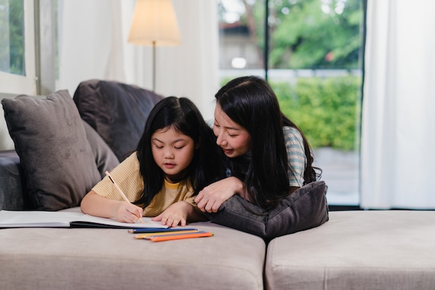 Asiatische Frau von mittlerem Alter unterrichten ihre Tochter, Hausarbeit zu tun und zu Hause zu zeichnen. Der glückliche Spaß der Lebensstilmutter und -kind verbringen Zeit zusammen im Wohnzimmer im modernen Haus am Abend.