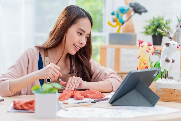 Asiatische Frauen verbringen den Wochenendtag für ihren Hobby-Tonskulpturen-Online-Kurs zu Hause, junge Erwachsene, die vom Tablet-Streaming-Kurs online in Schürzenkostümen lernen