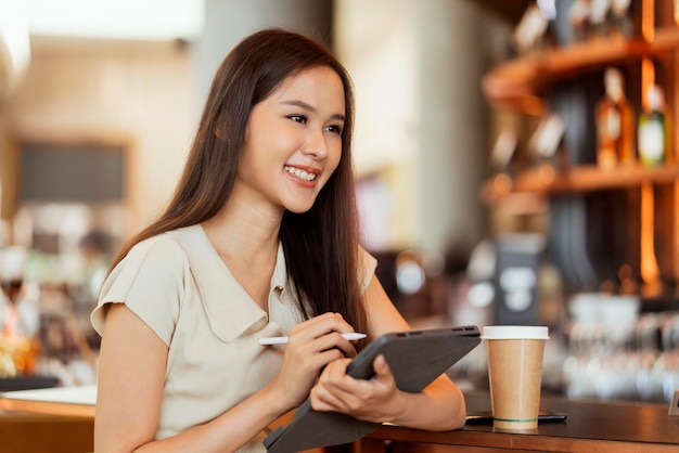 Asiatische Freiberuflerin, digitale Nomadenhand, die ein Tablet verwendet, das überall aus der Ferne arbeitet, sitzt an der Theke in der Nähe der Kaffeebar und kontaktiert den Kunden im Ausland mit einem fröhlich lächelnden Peacefel-Erfolgsabkommen