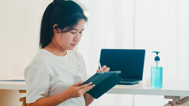 Asiatische freiberufliche Geschäftsfrau Freizeitkleidung unter Verwendung der Videokonferenz des Tablet-Arbeitsanrufs mit Kunden am Arbeitsplatz im Wohnzimmer von zu Hause aus, wenn soziale Distanzierung zu Hause bleibt und Selbstquarantänezeit.