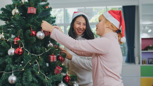 Asiatische Freundinnen verzieren Weihnachtsbaum am Weihnachtsfest. Das weibliche jugendlich glückliche Lächeln feiern Weihnachtswinterferien zusammen im Wohnzimmer zu Hause.