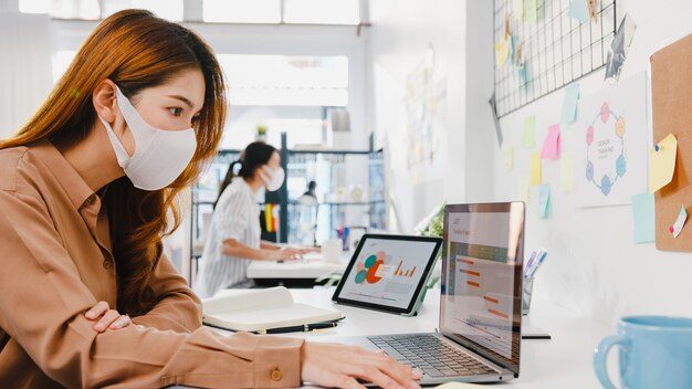 Asiatische Geschäftsfrau, die eine medizinische Gesichtsmaske zur sozialen Distanzierung in einer neuen normalen Situation zur Virusprävention trägt, während sie den Laptop bei der Arbeit im Büro verwendet.