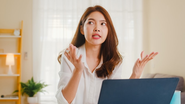 Asiatische Geschäftsfrau, die Laptop verwendet, spricht mit Kollegen über Plan in Videoanruf, während kluges Arbeiten von zu Hause im Wohnzimmer. Selbstisolation, soziale Distanzierung, Quarantäne zur Vorbeugung von Coronaviren.