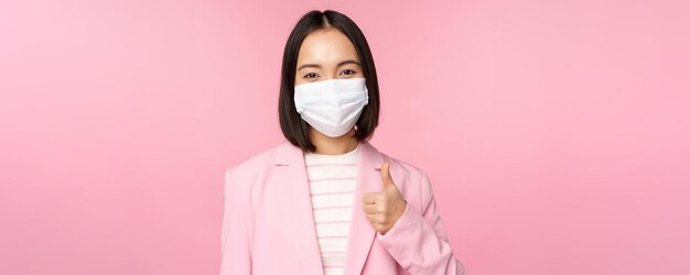 Asiatische geschäftsfrau in anzug und medizinischer gesichtsmaske zeigt daumen nach oben und empfiehlt das tragen persönlicher schutzausrüstung im büro während der covid19-pandemie auf rosa hintergrund