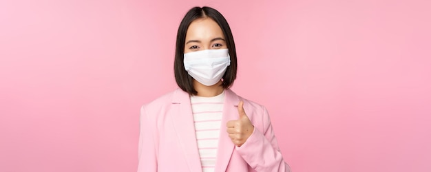 Asiatische geschäftsfrau in anzug und medizinischer gesichtsmaske zeigt daumen nach oben und empfiehlt das tragen persönlicher schutzausrüstung im büro während der covid19-pandemie auf rosa hintergrund