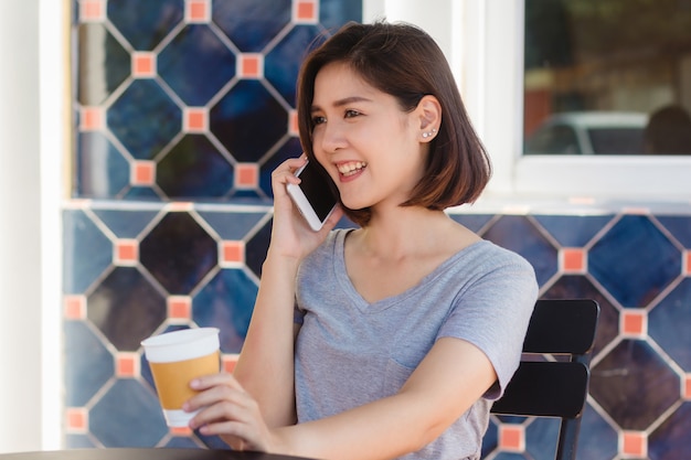 Asiatische Geschäftsfrauen des glücklichen Lächelns, die den Unterhaltungshandy sitzen im Café und im Halten einer Schale coffe verwenden