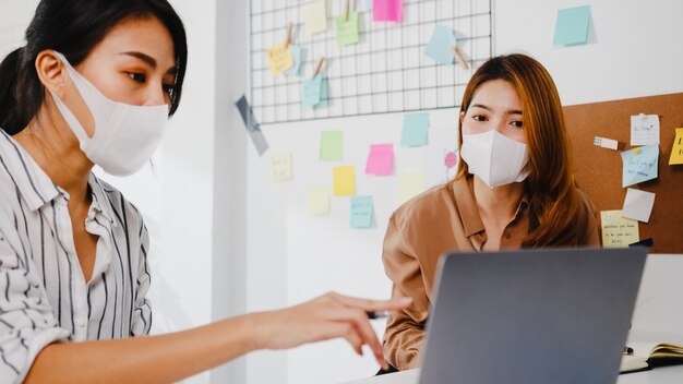 Asiatische Geschäftsleute, die Computerpräsentationen verwenden, Brainstorming-Ideen über neue Projektkollegen und tragen eine schützende Gesichtsmaske im neuen normalen Büro.