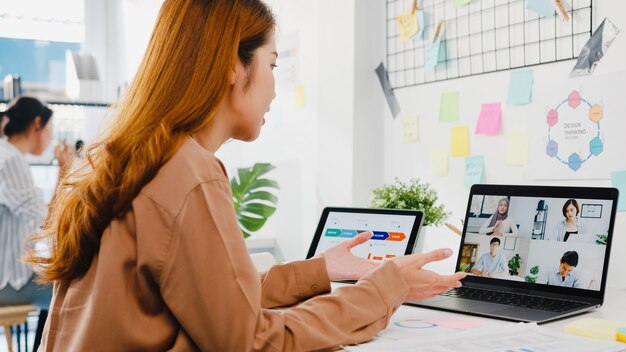 Asiatische Geschäftsleute, die Laptop verwenden, sprechen mit Kollegen, die ein Geschäfts-Brainstorming über den Plan in einem Videoanruf-Meeting in einem neuen normalen Büro diskutieren.