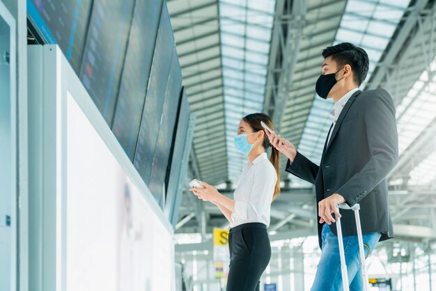Asiatische Geschäftsleute tragen einen Maskengesichtsschutz für Geschäftsreisen, um die Karte und den Flugplan am Informationsbildschirm im Terminalflughafen zu überprüfen
