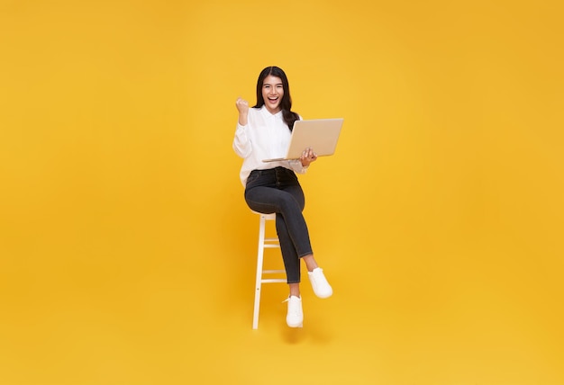 Asiatische glückliche lächelnde junge Frau, während sie Laptop benutzt, der auf weißem Stuhl sitzt