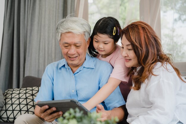 Asiatische Großeltern und Enkelin, die zu Hause Tablette verwendet. Der ältere glückliche Chinese, der Großvater und die Großmutter verbringen Familienzeit mit dem jungen Mädchen, das Social Media überprüft und liegen auf Sofa im Wohnzimmerkonzept