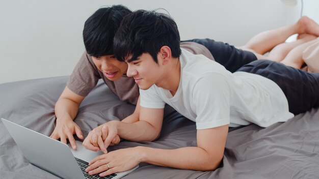 Asiatische homosexuelle lgbtq Mannpaare unter Verwendung des Computerlaptops am modernen Haus. Das junge männliche glückliche Asien-Liebhaber entspannen sich Rest zusammen, nachdem sie aufwachen und morgens den Film aufpassen, der auf Bett im Schlafzimmer am Haus liegt.