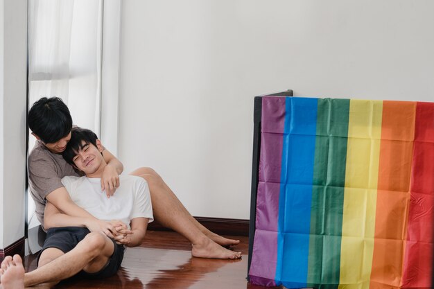 Asiatische homosexuelle Paare, die zu Hause auf dem Boden liegen und umarmen. Die jungen asiatischen LGBTQ + Männer, die glücklich küssen, entspannen sich Rest verbringen zusammen romantische Zeit im Wohnzimmer mit Regenbogenflagge am modernen Haus morgens.