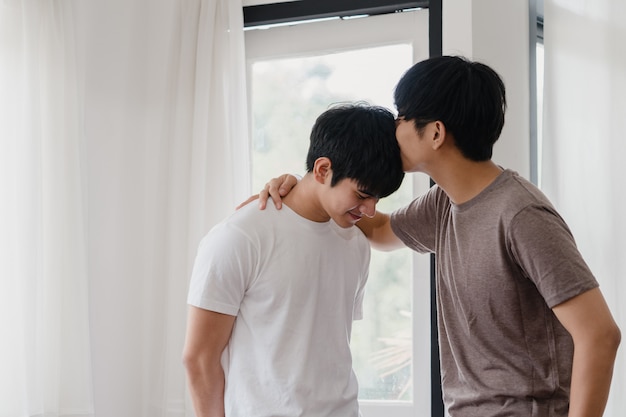 Asiatische homosexuelle Paare, die zu Hause nahe dem Fenster stehen und umarmen. Die jungen asiatischen LGBTQ + Männer, die glücklich küssen, entspannen sich Rest verbringen zusammen romantische Zeit im Wohnzimmer am modernen Haus morgens.