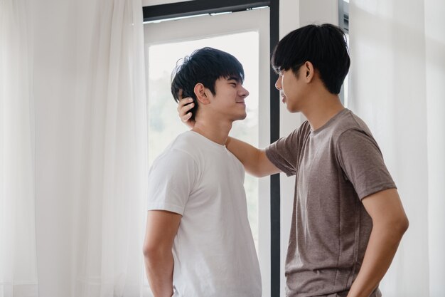 Asiatische homosexuelle Paare, die zu Hause nahe dem Fenster stehen und umarmen. Die jungen asiatischen LGBTQ + Männer, die glücklich küssen, entspannen sich Rest verbringen zusammen romantische Zeit im Wohnzimmer am modernen Haus morgens.