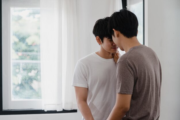 Asiatische homosexuelle Paare, die zu Hause nahe dem Fenster stehen und umarmen. Die jungen asiatischen LGBTQ + Männer, die glücklich küssen, entspannen sich Rest verbringen zusammen romantische Zeit im Wohnzimmer am modernen Haus morgens.